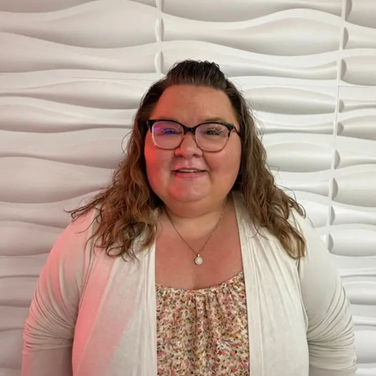Portrait of a smiling woman with glasses, wearing a floral top and light cardigan, standing against a white wave-textured wall. - My Topp Home