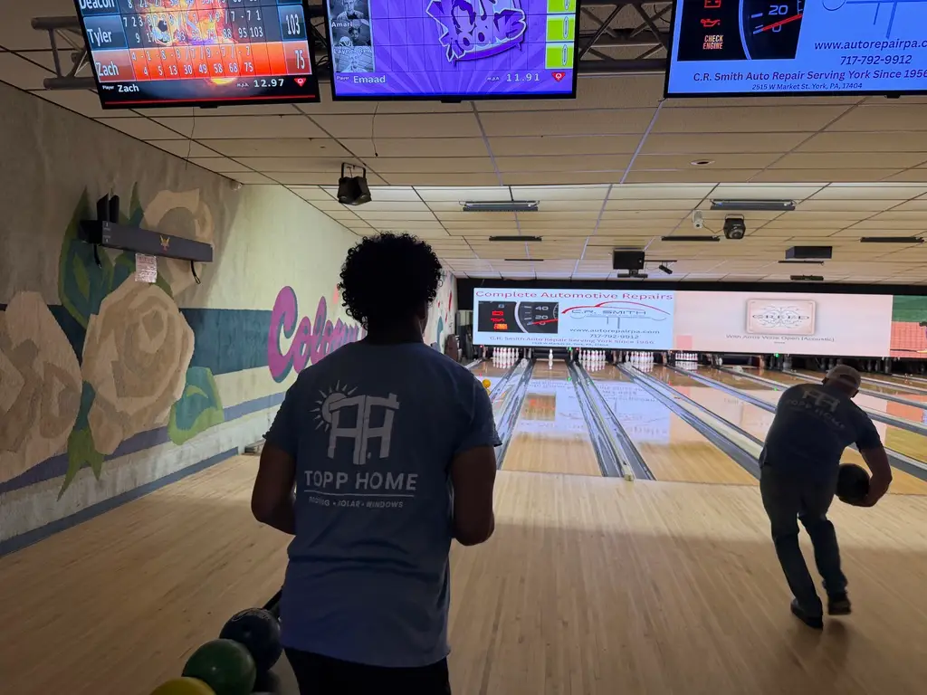 Bowling alley scene with a player in a blue shirt standing near the lanes, murals on the wall and digital scoreboards overhead.