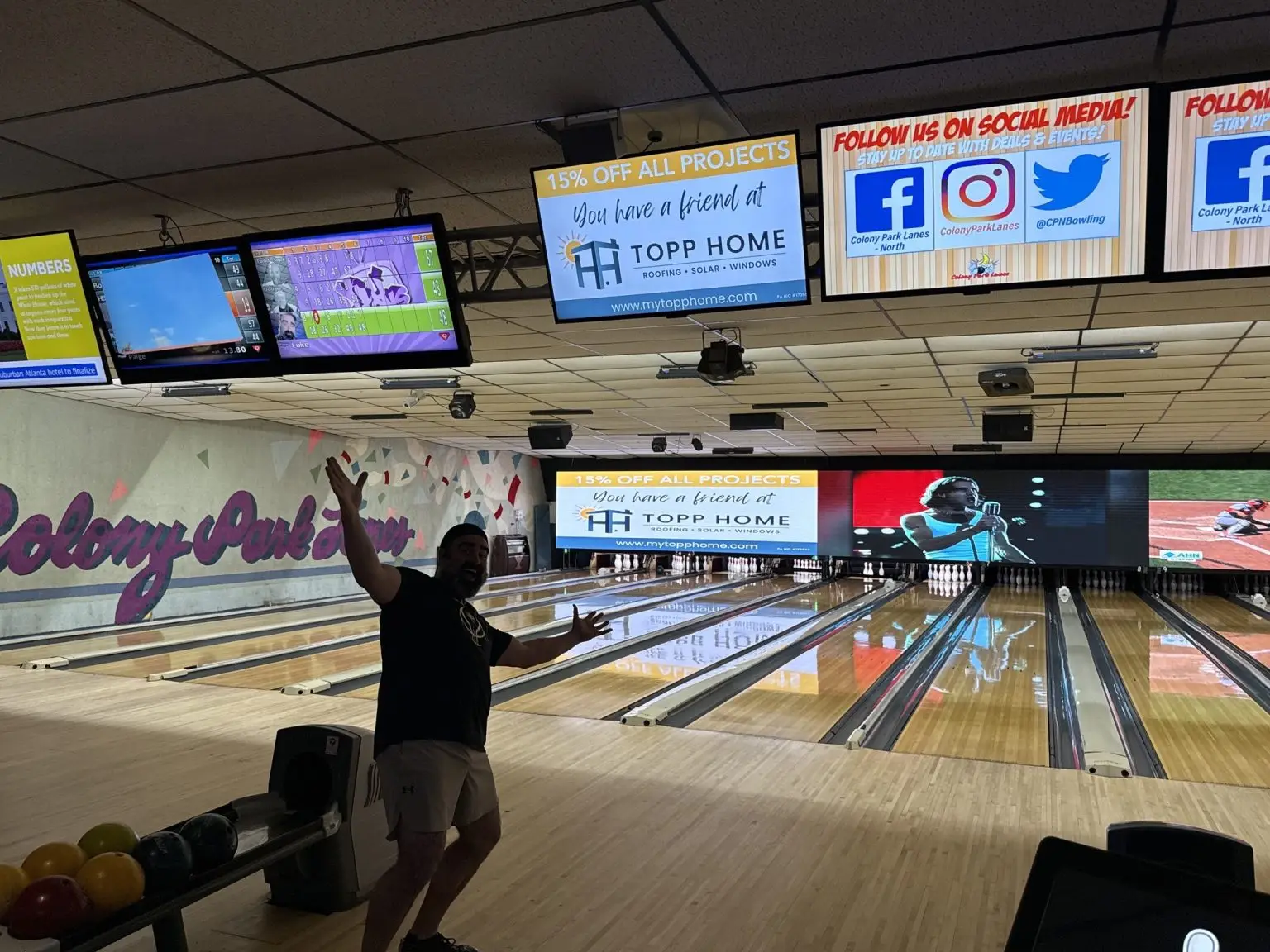 Man in a dark shirt and shorts raises arms in celebration at a bowling lane, with colorful balls on the rack nearby and bright score screens above.