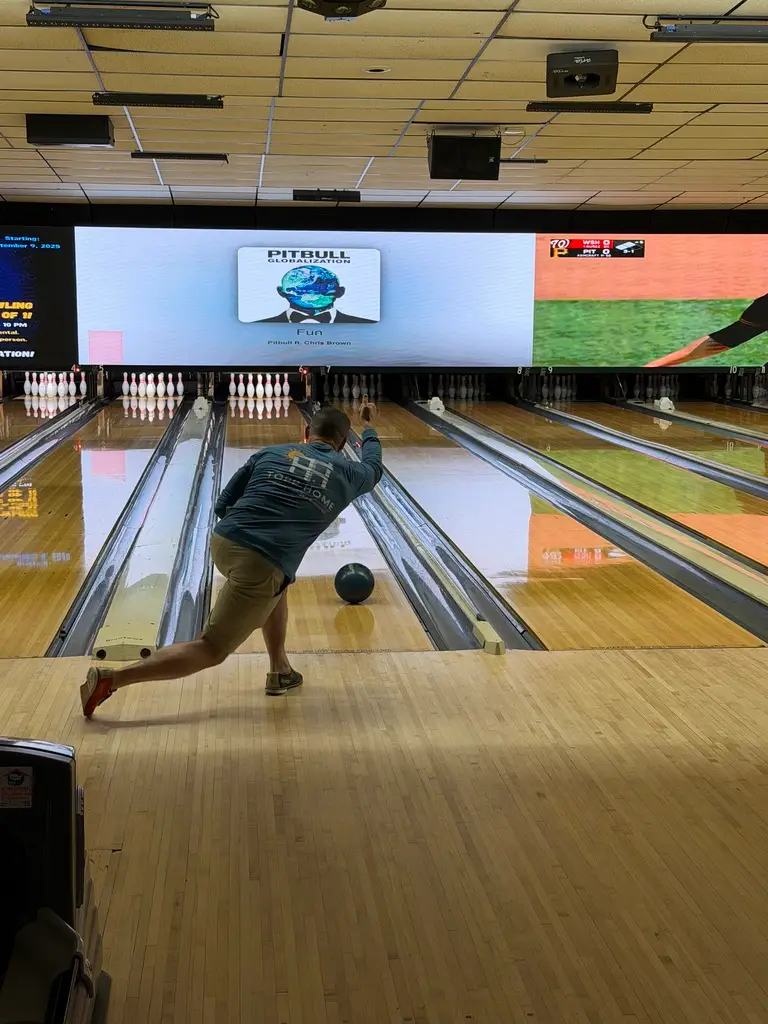 Bowler in a teal shirt releases a ball down a wooden bowling lane toward the pins at an indoor bowling alley.