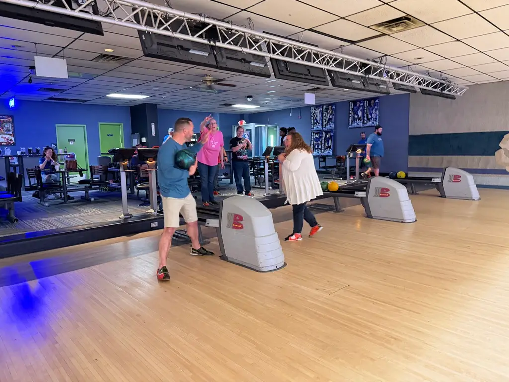 Group of people bowling and socializing at a bowling alley, with lanes, ball returns, and overhead lighting visible in the background.
