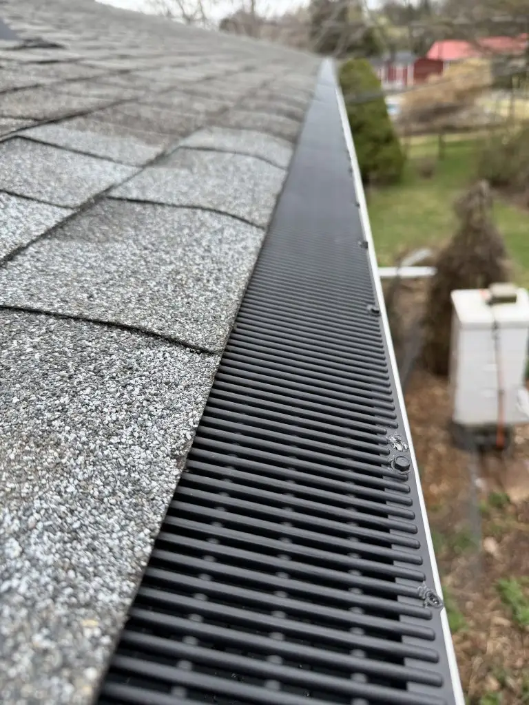 Close-up of a roof edge with gray asphalt shingles and a black vented gutter along the eave; backyard is blurred in the background. - My Topp Home