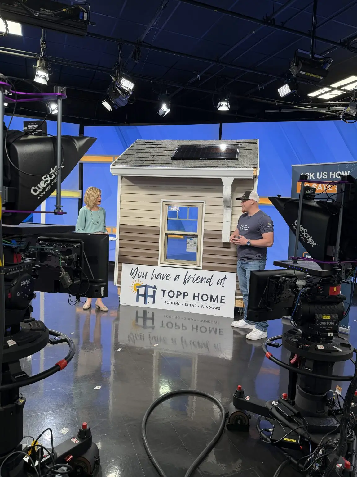 Studio set with a small beige house prop and two hosts talking, surrounded by overhead lights and cameras on a blue backdrop.
