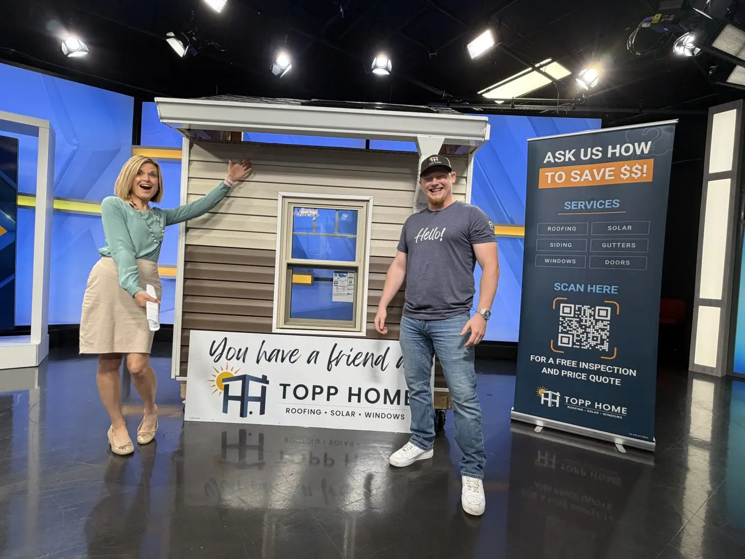 Two smiling people pose on a TV studio set next to a house display and a tall promo banner reading 'ASK US HOW TO SAVE $$' and 'Topp Home' signage on the barrier.