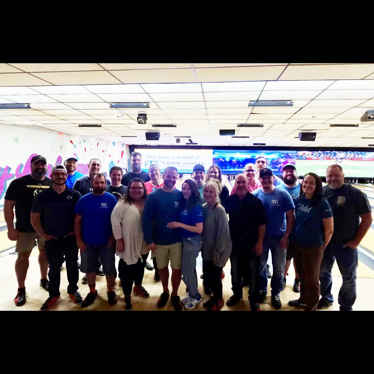 Group of about 20 coworkers posing for a photo in a bowling alley, with lanes and a mural wall in the background.