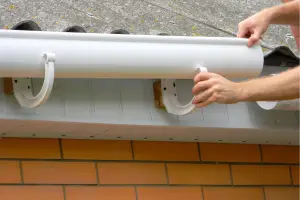 Person attaches a white rain gutter pipe to the roof gutter brackets under a roof edge on a brick house. - My Topp Home