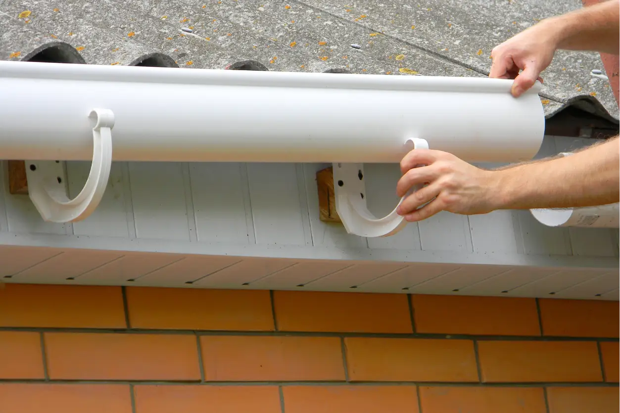 Person attaches a white rain gutter pipe to the roof gutter brackets under a roof edge on a brick house. - My Topp Home