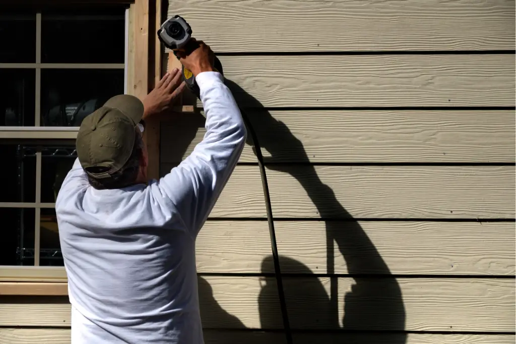 Replaced or Cleaned - My Topp Home Man in a white long-sleeve shirt and olive cap uses a power drill to fasten wooden siding beside a window on a house; strong sunlight casts a long shadow on the wall. - My Topp Home