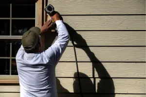 Man in a white long-sleeve shirt and olive cap uses a power drill to fasten wooden siding beside a window on a house; strong sunlight casts a long shadow on the wall. - My Topp Home - My Topp Home